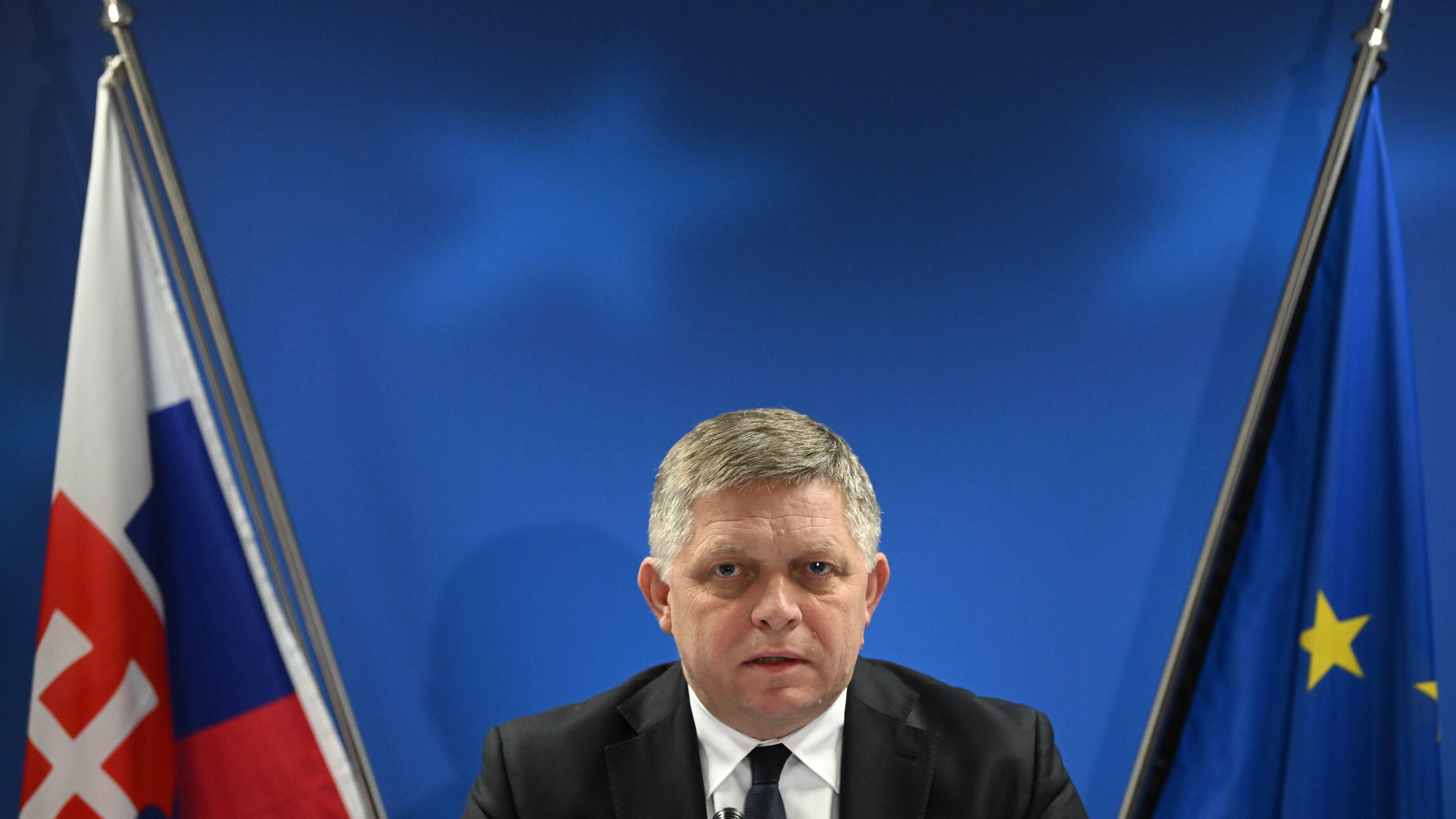 Robert Fico sitzt zwischen der slowakischen und der europäischen Flagge und schaut in die Kamera