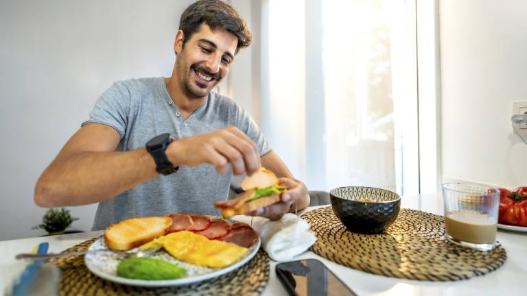 Ein Mann sitzt in der Küche am Frühstückstisch. Vor ihm stehen verschiedene Lebensmittel wie Obst, Brot, Müsli. Zu trinken hat er einen Milchkaffee. Der Mann belegt sich gerade ein Brot. Sein Handy liegt neben dem Teller.