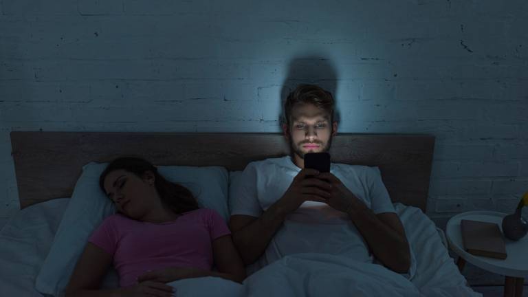 Ein Pärchen liegt im Bett. Der Mann kann nicht schlafen und schaut auf sein Handy. Das Smartphone-Bildschirmlicht hält ihn weiter vom Schlafen ab.