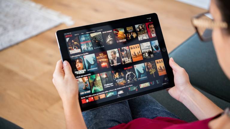 Eine Person sitzt auf dem Sofa und hält ein Tablet in der Hand. Sie ist damit beschäftigt, einen Film oder eine Serie auszuwählen.