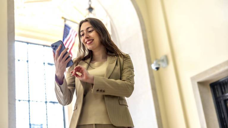 Hellhäutige US-Amerikanerin trägt einem braunen Anzug und lächelt, während sie mit einem Smartphone in einem hellen, gewölbten Gebäude Dating-Profile checkt. Über ihr - in dem Gebäude in Manhattan – ist die US-amerikanische Flagge gehisst.