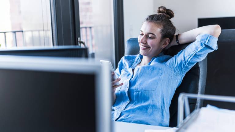 Eine Frau sitzt im Büro am Schreibtisch, Auf ihrem Schreibtisch stehen Computer und Akten. Sie nimmt sich einen Moment Zeit, um zu entspannen und blickt auf ihr Smartphone.