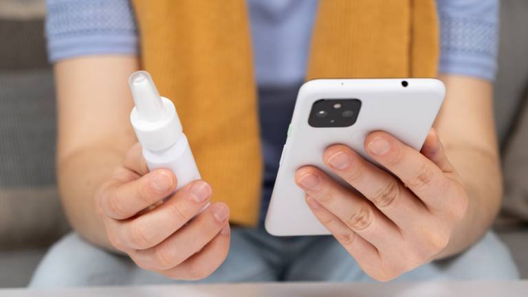 Eine Person hält in der rechten Hand ein Schnupfen-Spray, in der rechten Hand das Smartphone, um Arzt um Krankschreibung zu bitten.