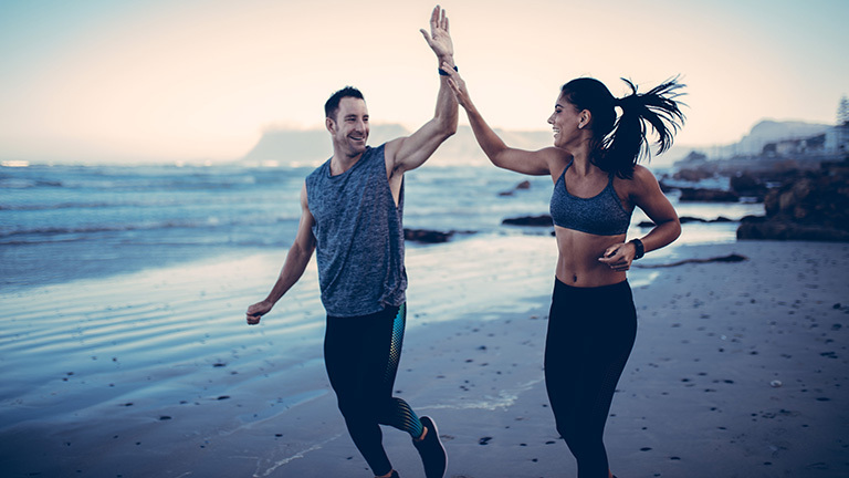 Zwei Sportler spornen sich beim Jogging an einer Küste durch lächeln und sich aufeinander zubewegenden Handbewegungen an