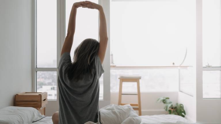 Eine Frau streckt sich, nachdem sie aufgewacht ist. Sie blickt aus den Fenstern aus ihrem Bett. Der Tag hat begonnen.