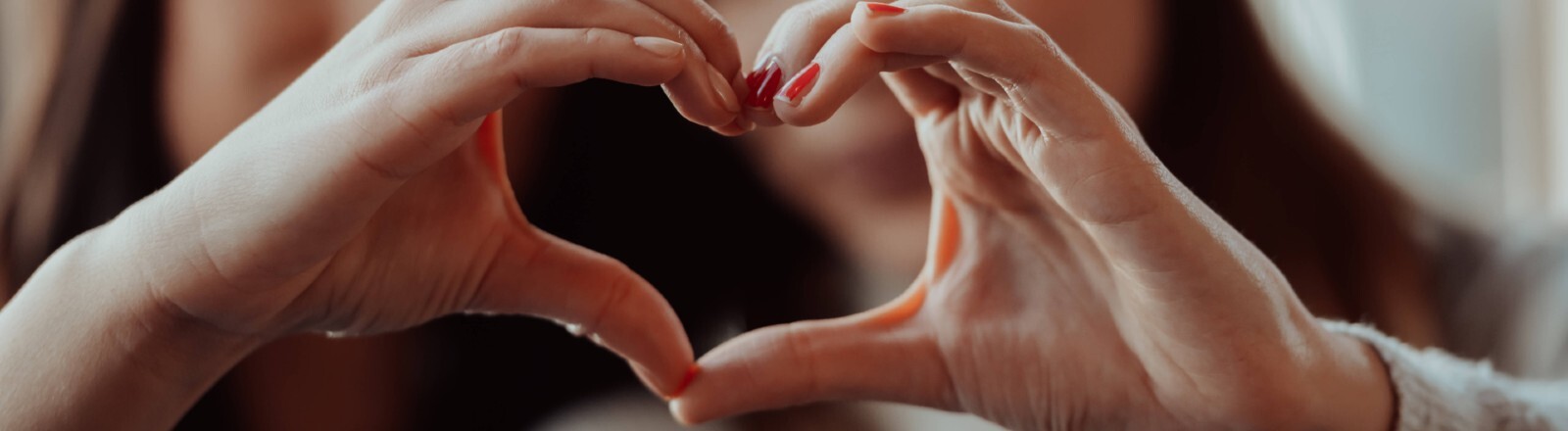Zwei Frauen halten ihre Hände so zusammen, dass sie gemeinsam ein Herz ergeben. Die Herz-Hand ist im Vordergrund. Die beiden Frauen sind unscharf im Hintergrund.