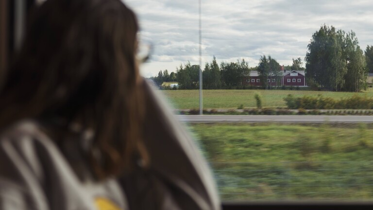Eine junge Frau sitzt im Zug und schaut aus dem Fester raus. Draußen sieht man grüne Landschaften.