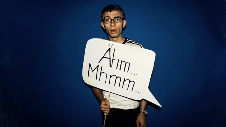 Mann mit Brille hält Schild hoch mit der Aufschrift Ähm... Mhmm...