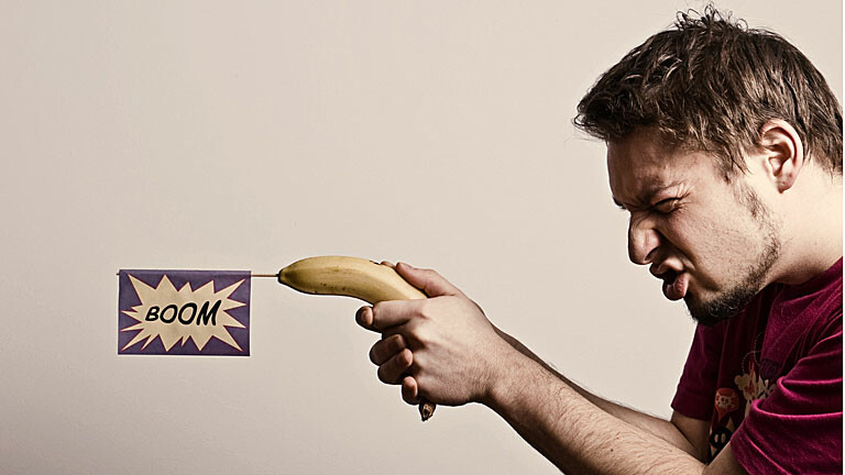 Mann "schießt" mit Banane, in der Schild mit Aufschrift "Boom" steckt