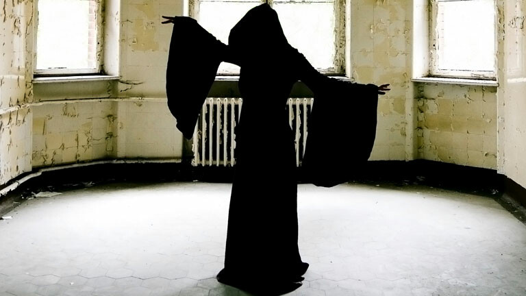 Eine Frau in einem schwarzen Umhang hebt beschwörend die Arme.