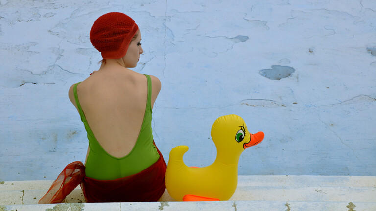 Ein Frau sitzt in Badeanzung und Bademütze neben einem großen Gummiente an einem ausgetrockneten Becken.