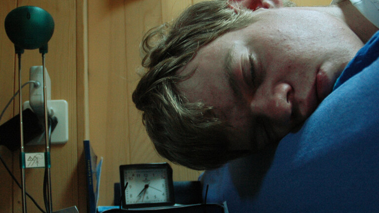 Ein junger Mann schläft komatös neben seinem Wecker.