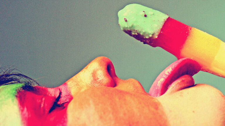 Eine Frau leckt an einem Eis, das grün, rot, orange und gelb ist. Ihr Gesicht ist in denselben Farben angemalt. Lediglich ihr Gesicht im Profil und das Eis sind zu sehen.