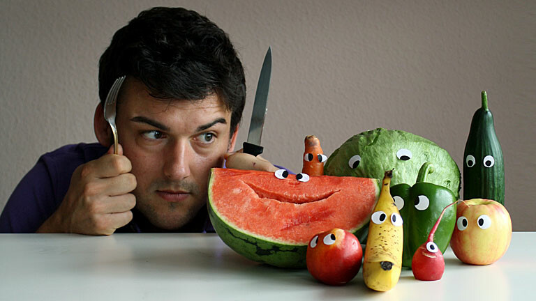 Ein Mann, Messer und Gabel gezückt, starrt auf Gemüse & Obst mit angeklebten Augen