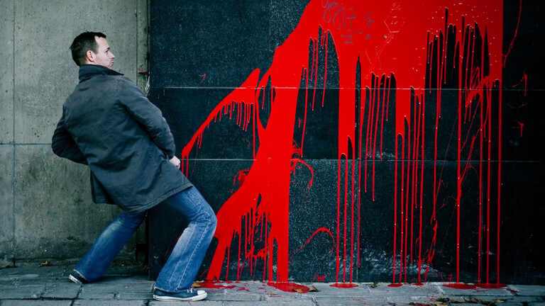 Ein Mann steht an einer Wand, als ob er pinkeln würde. Auf der Wand verläuft rote Farbe.