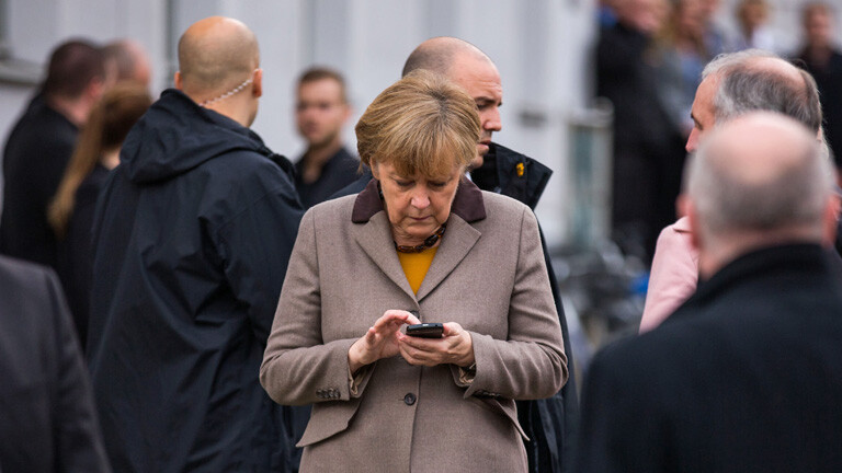 Kanzlerin Angela Merkel tippt eine SMS auf ihrem Mobiltelefon, drumherum stehen Leute (03.11.2014); Bild: dpa