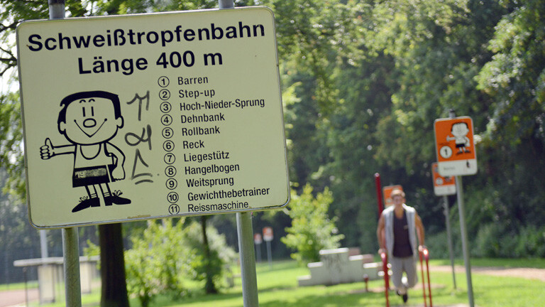 Deutschlands erster Trimm-Dich-Pfad. Auf einem Schild ist die Zeichnung eines Männchen, das den Daumen nach oben streckt. Darüber steht "Schweißtropfenbahn"; Bild: dpa