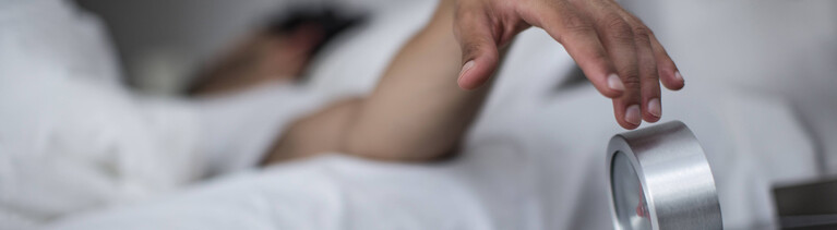 Ein Mann liegt im Bett und greift herüber zu seinem Wecker, um den Alarm auszuschalten.