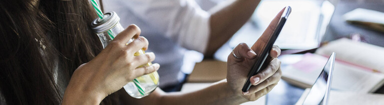 Frau vor dem Rechner, mit Limonade in der einen und Smartphone in der anderen Hand.