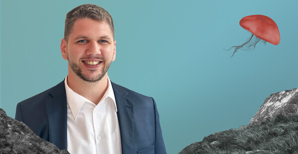 Ein Mann mit kurzen, grauen Haaren steht lächelnd vor einem blauen Hintergrund. Im Hintergrund Berge und Quallen.