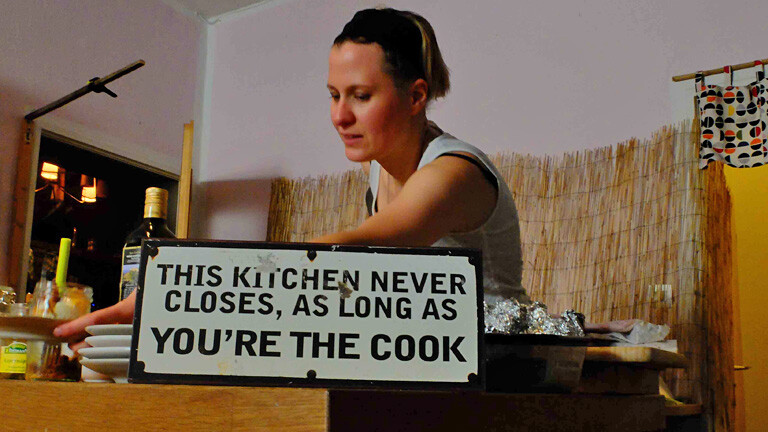 Eine Frau steht hinter einem Küchenbord. Auf diesem steht ein Schild mit dem Schriftzug: this kitchen never closes, as long as you're the cook.