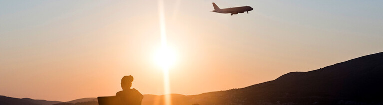 Eine Frau sitzt mit Laptop am Strand im Hintergrund ein Flugzeug.