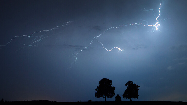 Zwei Bäume vor düsterem Gewitterhimmel mit Blitz.