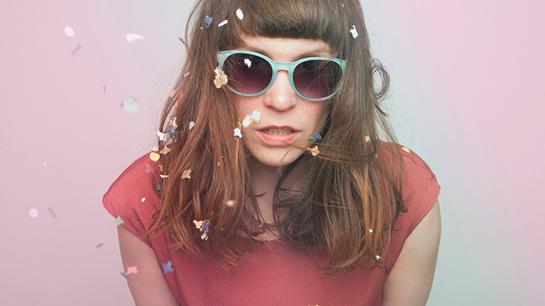 Eine Frau schaut schlecht gelaunt in die Kamera; sie trägt eine Sonnenbrille; Konfetti rieselt auf sie nieder.