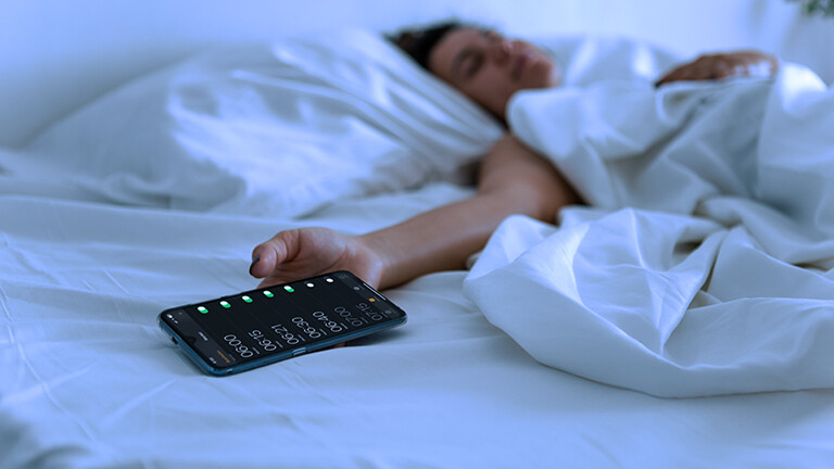 Eine Frau liegt mit ausgestrecktem Arm auf dem Bett und hält ein Smartphone in der Hand, auf dem viele Weckzeiten eingestellt sind.