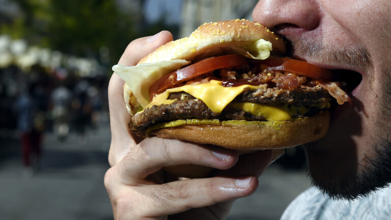 Ein bärtiger Mann beißt in einen Hamburger. Sein Mund ist weit geöffnet, der Hamburger ist üppig belegt. Eine Käsescheibe, eine Tomatenscheibe und Salat sind zu sehen. Auch mehrere Frikadellen sind zwischen den Brötchenhälften.