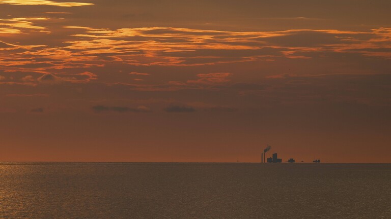Sonnenlicht spiegelt sich auf dem Meer. Am Horizont ist die Silhouette einer Fabrik zu sehen