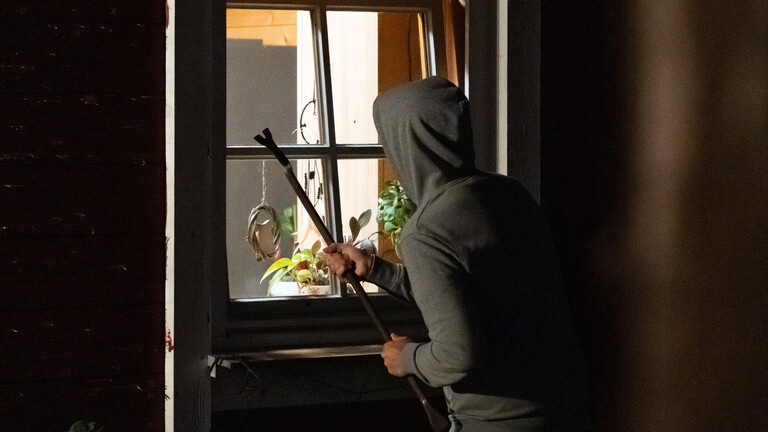 Symbolbild: Ein Einbrecher mit Kapuze und Brecheisen steht nachts vor einem gekippten Fenster.