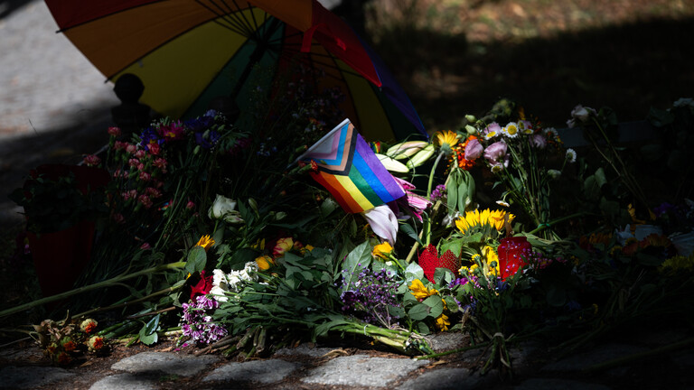 Die Sonne scheint auf Blumen und eine Progress-Pride-Fahne, die zum Gedenken an die Opfer des Anschlags auf den CSD abgelegt wurden.