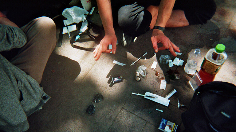 Analoge Fotografie aus dem Projekt "(Über)Leben im Risikoumfeld" zweier nicht erkennbarer Menschen, die auf dem Boden sitzend Drogenkonsum vorbereiten. Man sieht Knie und ein Paar Hände, auf dem Pflasterstein liegen unter anderem drei Einwegspritzen, eine Sonnenbrille, eine Zigarettenschachtel und Tütchen mit Drogen, daneben stehen Getränkeflaschen.