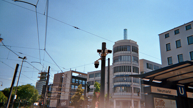 Analoge, weitwinklige Fotografie aus dem Projekt "(Über)Leben im Risikoumfeld", dass die oberen Etagen moderner Gebäude am Kölner Neumarkt vor blauem, von Straßenbahnkabeln durchzogenen Himmel zeigt. In der Mitte des Bildes steht ein Mast, an dem drei Überwachungskameras angebracht sind. Am rechten unteren Rand des Bildes ist an einem Wartehäuschen das blaue Zeichen U für U-Bahn und ein Schild "Neumarkt" zu sehen.