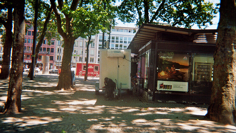 Analoge Fotografie aus dem Projekt "(Über)Leben im Risikoumfeld": Ein Mensch kauert hinter einem Kiosk am Kölner Neumarkt.