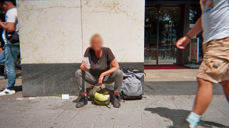 Analoge Fotografie aus dem Projekt "(Über)Leben im Risikoumfeld", die eine Person zeigt, die am Rande einer Fußgängerzone auf dem Boden sitzenden bettelt, das Gesicht ist unkenntlich gemacht.
