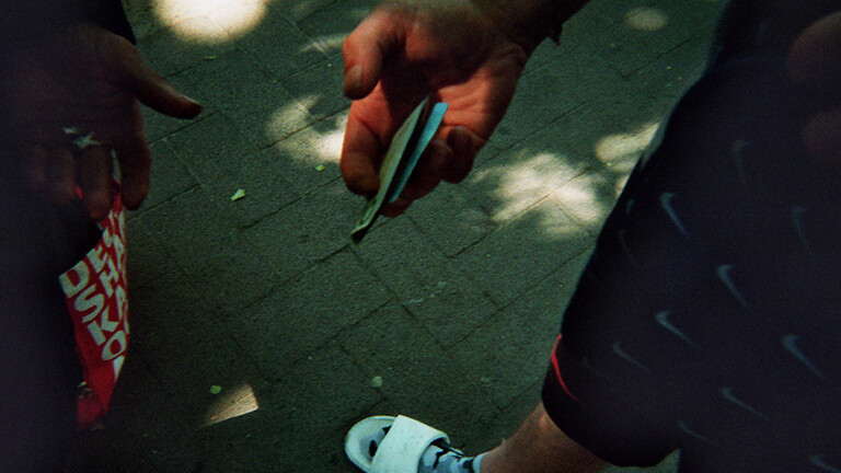 Analoge Fotografie aus dem Projekt "(Über)Leben im Risikoumfeld": Auf dem Bild , das Richtung Boden fotografiert ist, sieht man zwei Hände, die eine reicht der anderen gefaltete Geldscheine. Am Boden ist ein Fuß in weißen Badeschlappen erkennbar.