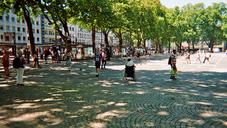Eine Analoge Fotografie aus dem Projekt "(Über)Leben im Risikoumfeld" zeigt eine Totale des Kölner Neumarkts an einem sonnigen Tag mit Passanten.