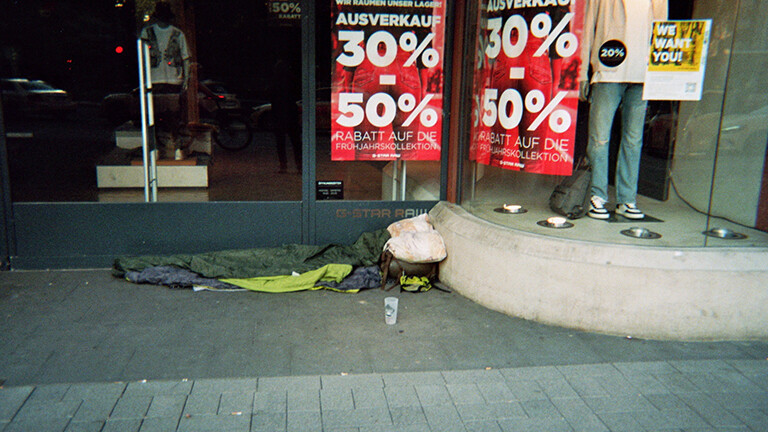 Analoge Fotografie aus dem Projekt "(Über)Leben im Risikoumfeld": Das Foto zeigt einen leeren Schlafplatz mit Schlafsack, einem Kopfkissen auf einem Rucksack und einem leeren Plastikbecher zum Betteln davor auf dem blanken Pflasterboden vor dem Schaufenster eines Bekleidungsgeschäfts.