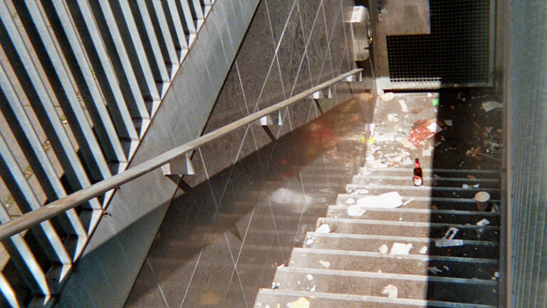 Analoge Fotografie aus dem Projekt "(Über)Leben im Risikoumfeld": Blick eine Treppe hinab zu einer verschlossenen Tür in einem Untergeschoss. Die Sonne scheint halb in den Schaft und beleuchtet viel herumliegenden Müll und eine geöffnete Kölschflasche.
