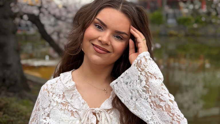 Protagonistin Bella steht im Park vor einem Baum und lächelt in die Kamera.