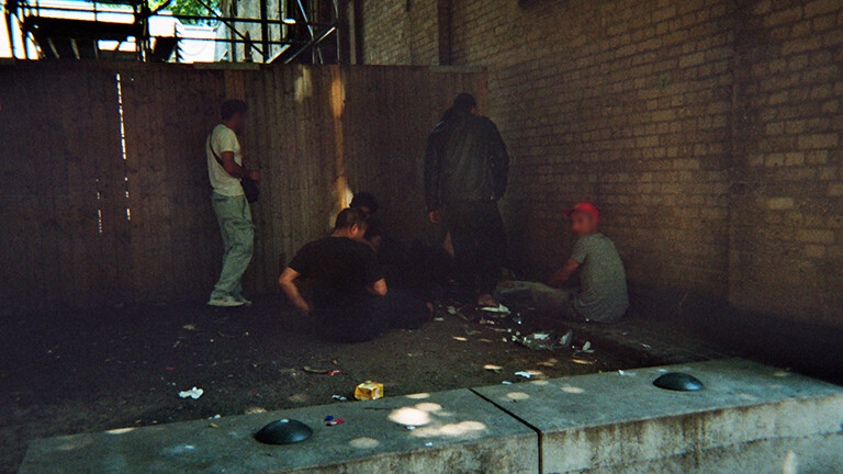 Analoge Fotografie aus dem Projekt "(Über)Leben im Risikoumfeld" einer Gruppe nicht erkennbarer Menschen, die in einer schattigen Ecke zwischen einer Hauswand und einer Baustellenwand inmitten von Müll stehen oder sitzen und offenbar gemeinsam Drogen konsumieren