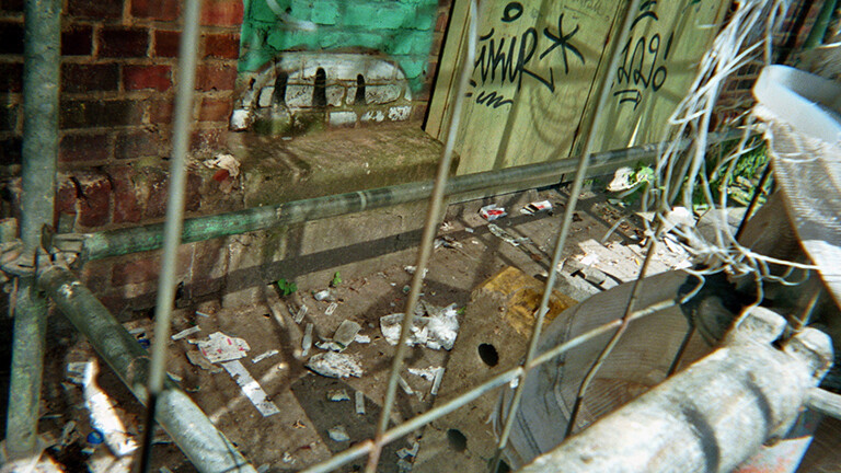 Analoge Fotografie aus dem Projekt "(Über)Leben im Risikoumfeld":  Hinter einem Bauzaun unter einem Baugerüst an der mit Tags bemalten Wand eines Industriegebäudes liegt viel Müll und leere Drogentütchen