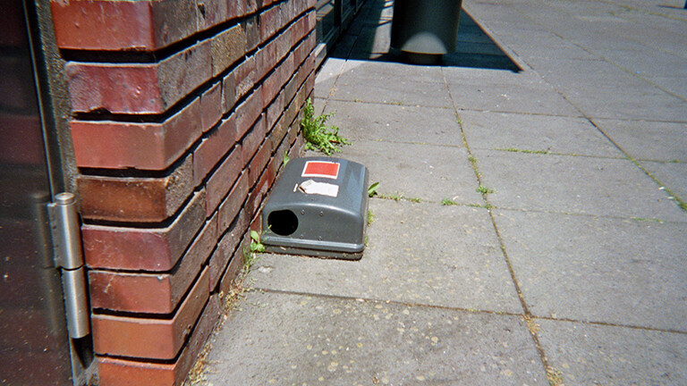 Analoge Fotografie aus dem Projekt "(Über)Leben im Risikoumfeld" einer Plastikrattenfalle, die an einer Backsteinwand auf einem Steinboden aufgestellt ist.