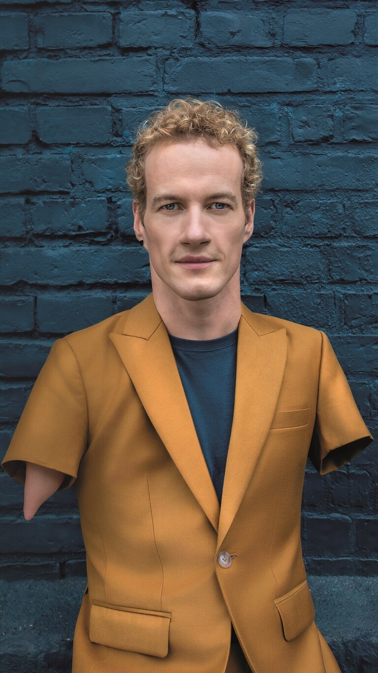 Porträt von Janis in einem senfgelben Sakko vor einer dunkelblauen Backsteinwand