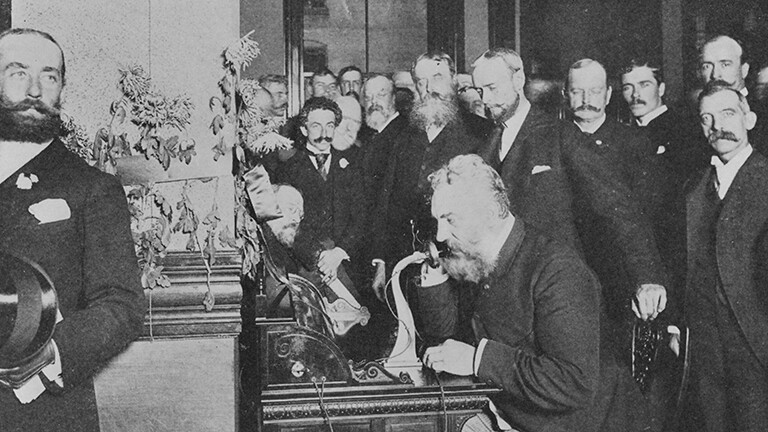 Schwarz-weißes Archivfoto: Alexander Graham Bell sitzt an einem Telefonapparat 1892, um ihn herum steht eine Gruppe Männer.