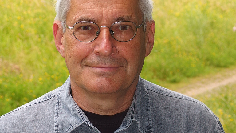 Portrait von Jürgen Heinze. Er steht draußen, im Hintergrund ist eine Wiese. Er trägt ein graues Hemd, eine Brille und blickt direkt in die Kamera.