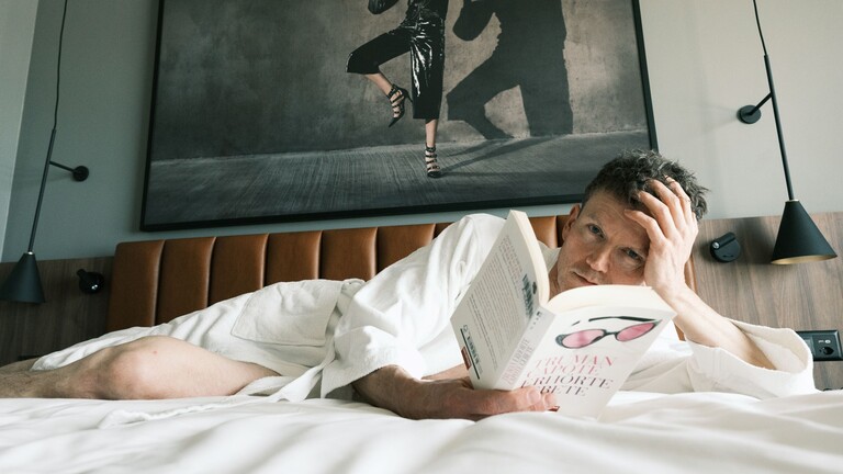 Noah trägt einen Bademantel und liegt im Bett, er liest ein Buch