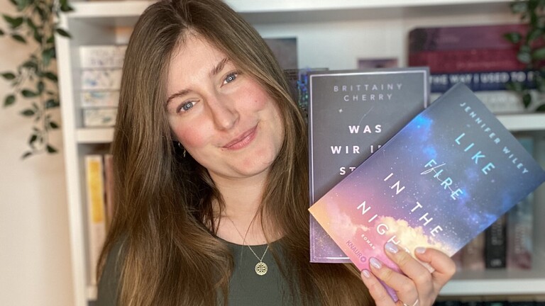 Benita steht vor ihrem Bücherregal, hält zwei Bücher einer Reihe in der Hand und lächelt in die Kamera.